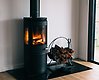 En mindre svart kamin där det lyser eldslågor inuti. Till höger om kaminen står en rund korg med ved.