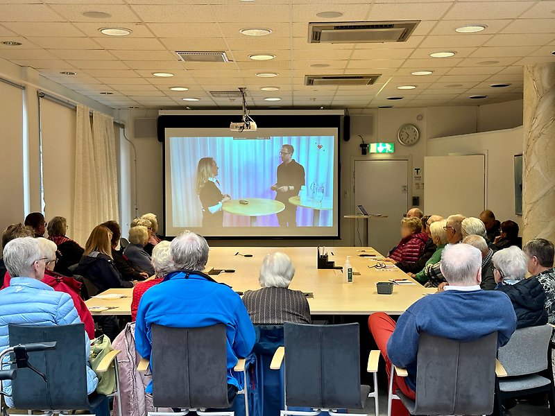 I en sammanträdeslokal sitter många äldre personer runt ett stort bord och ser på en webbföreläsning som sänds på en projektorduk.