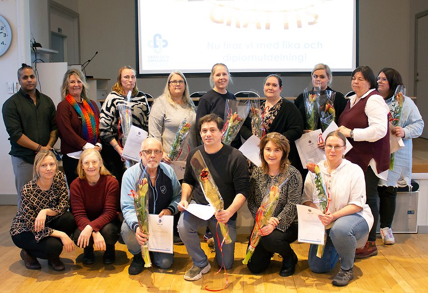 Gruppbild på femton personer på två rader, den undre raden står på knä. Alla håller diplom och blommor i händerna. På en projektorduk i bakgrunden står det "Grattis! Nu firar vi med fika och diplomutbildning!"