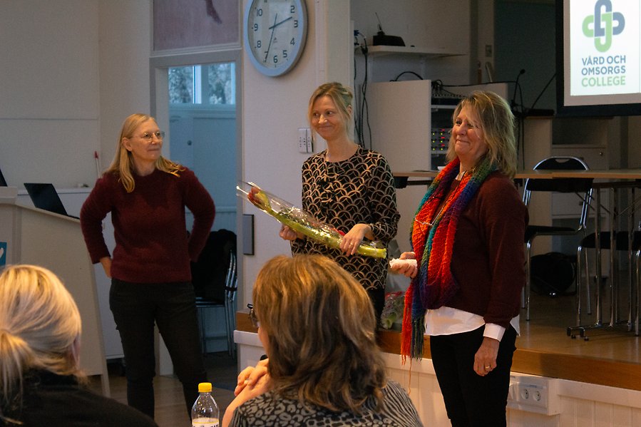 Tre kvinnor som står framför personer sittande vid bord.
