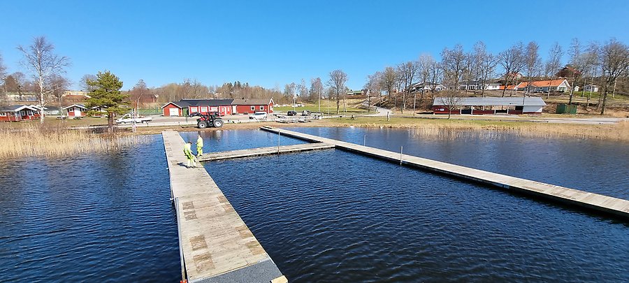I strålande solsken går två personer i arbetskläder på en stor flytbrygga. Bryggan är byggd som ett H. Himmel och vatten är klarblått, vattnet mörkare blå. Vid stranden syns vass och längre in på land en dansbana och klubbhusbyggnader.