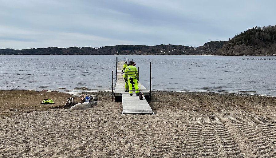 På en lång brygga i en fjord går två personer i arbetskläder. Himlen och vattnet är gråblått. På sandstranden syns hjulspår efter en traktor. Vid horisonten böljar låga berg.