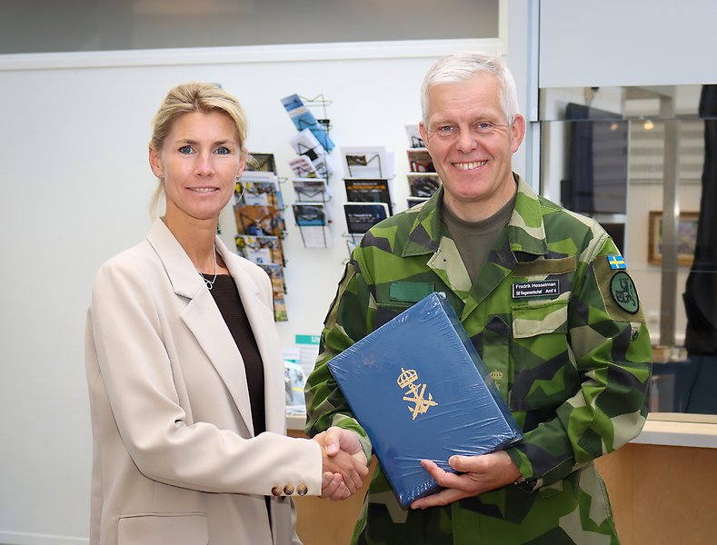 Två personer skakar hand varav en har militäruniform och en bok i handen