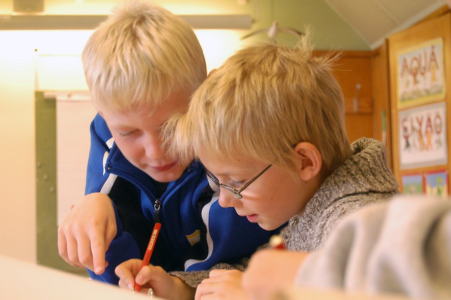 Två pojkar sitter respektive står vid en skolbänk. Pojken som står hjälper den andra pojken.
