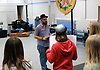 Barn står på scenen och en ser deras ryggar eller bakhuvuden. En av dem håller i ett manus. Framför dem står en kulturpedagog och håller i ett manus. I bakgrunden syns olika musikinstrument.