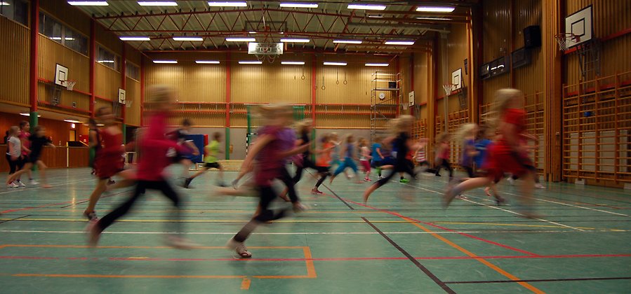 Foto I en idrottshall. Där springer många barn åt höger. De leker kom alla mina kycklingar. De springer så fort att de blir suddiga på fotot.
