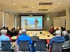 I en sammanträdeslokal sitter många äldre personer runt ett stort bord och ser på en webbföreläsning som sänds på en projektorduk.