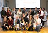 Gruppbild på femton personer på två rader, den undre raden står på knä. Alla håller diplom och blommor i händerna. På en projektorduk i bakgrunden står det "Grattis! Nu firar vi med fika och diplomutbildning!"
