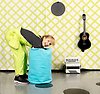 En glad kvinna i limegröna snickarbyxor sträcker sig ned i en turkos säck. I bakgrunden på den snedrutiga tapeten hänger en gitarr. På golvet står ett dragspel.