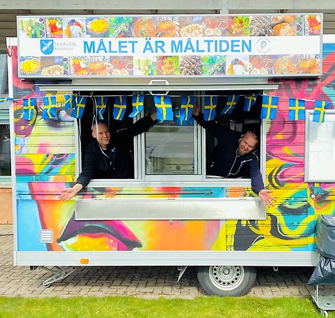 En foodtruck målad i glada färger. Två personer sträcker glatt ut sina armar ur luckorna. Längs med vagnen hänger vimplar med svenska flaggor. På en skylt står det Målet är måltiden tillsammans med Munkedals kommuns och Foodfluencers logotyper.