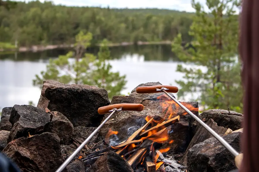 Korvar som grillas på grillspett över en öppen eld. I bakgrunden syns en sjö.