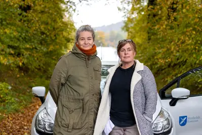Två kvinnor står framför en vit bil utomhus bland färgstarka träd en höstdag.